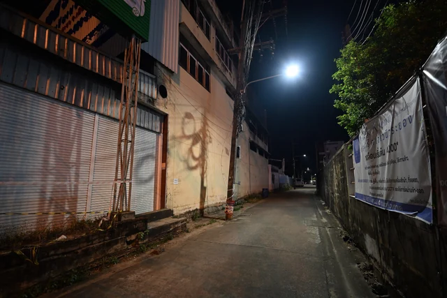 Gece vakti dar bir sokak; solda kepenkleri kapalı bir dükkân, önünde ahşap destekler ve şerit, uzakta tek bir sokak lambası yanıyor.
